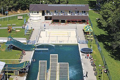 Celkový pohled na areál Acrobat Park. V popředí jsou odrazové plochy můstků, dále bazén, hřiště na plážový volejbal, v pozadí objekt s restaurací, ubytovací kapacitou, šatnami, technickým a administrativním zázemím. Objekt je vybaven střešní krytinou Ruukki