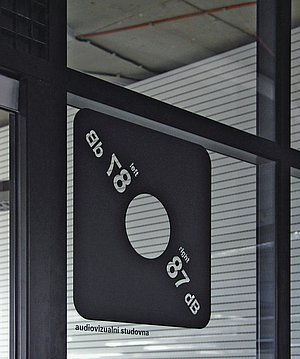 Piktogram audiovizuální studovny vyjádřený hodnotou 87 dB (maximální povolená hladina hluku na pracovišti při plném úvazku)