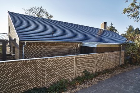 Pohled na dům z ulice: pultová střecha tvořená černou betonovou krytinou, plot obložený stejnými cihlami jako sám dům