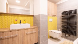 I v koupelně zůstalo zachováno stejné barevné ladění – drobná žlutá a bílá kruhová mozaika kontrastuje s velkými formáty šedých dlaždic na podlaze